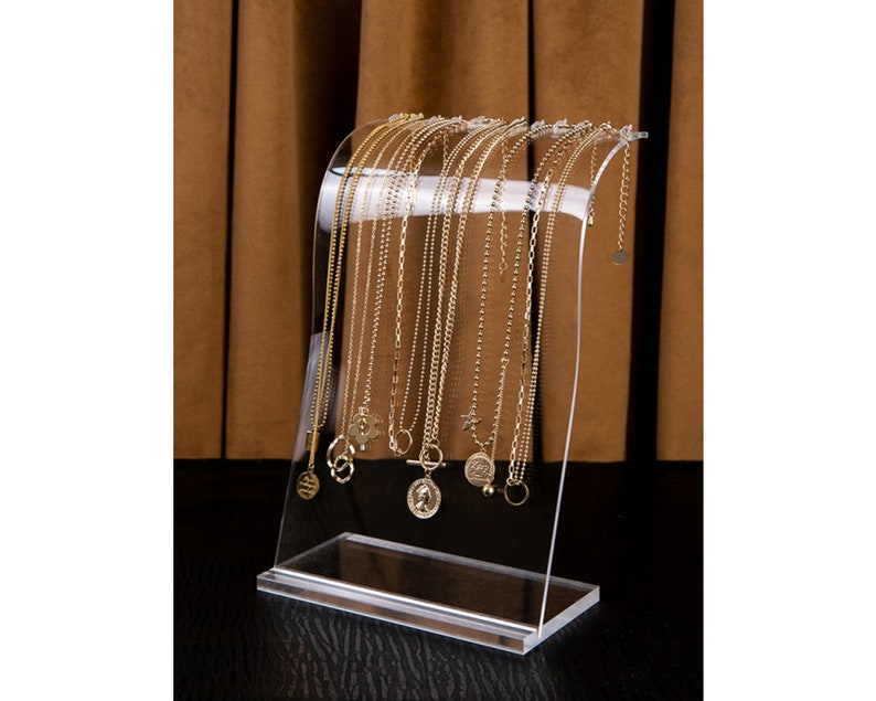 Peut inclure: Pr&eacute;sentoir &agrave; colliers en acrylique transparent pr&eacute;sentant plusieurs colliers dor&eacute;s avec diff&eacute;rents mod&egrave;les de pendentifs. Le pr&eacute;sentoir a un design incurv&eacute; et une base rectangulaire, id&eacute;al pour les bijoux.