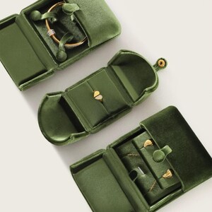 May include: Three olive green velvet jewelry boxes with gold-tone snap closures. The top box holds a gold bangle, the middle box contains a gold heart-shaped ring, and the bottom box displays a gold chain necklace with two small gold stud earrings.