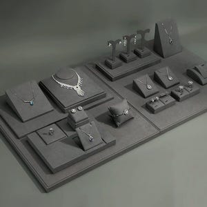 May include: A display of jewelry on gray velvet platforms. The display includes necklaces, earrings, rings, and bracelets. The jewelry is silver-toned with clear gemstones. The arrangement is on a large, rectangular gray surface.