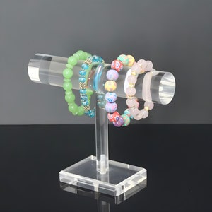 Puede incluir: Un expositor de pulseras de acrílico transparente con cuatro pulseras de cuentas de colores. El expositor tiene una varilla redonda y una base rectangular.