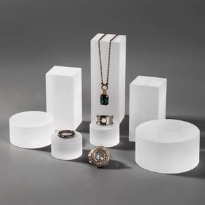 Könnte beinhalten: Schmuckpräsentation mit Ringen und einer Halskette mit Anhänger. Der Schmuck wird auf weißen geometrischen Podesten unterschiedlicher Formen und Größen präsentiert. Der Hintergrund ist einfarbig grau.