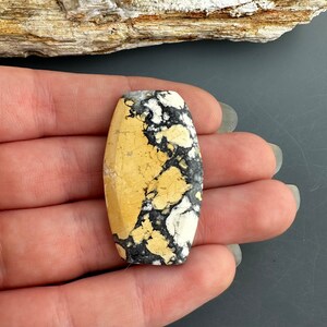 Puede incluir: Una piedra de cabuj&oacute;n de forma ovalada con un patr&oacute;n amarillo, negro y blanco. La piedra es lisa y pulida, con una apariencia marmoleada &uacute;nica.