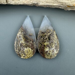 May include: Pair of teardrop-shaped agate cabochons with a white, gray, and brown dendritic pattern. The cabochons are translucent and have a smooth, polished surface.