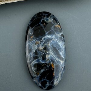May include: Oval-shaped gemstone with a polished surface. The stone features a dark blue and black base with swirling patterns of white and gold. A small hole is drilled at the top. The gemstone is set against a gray background.