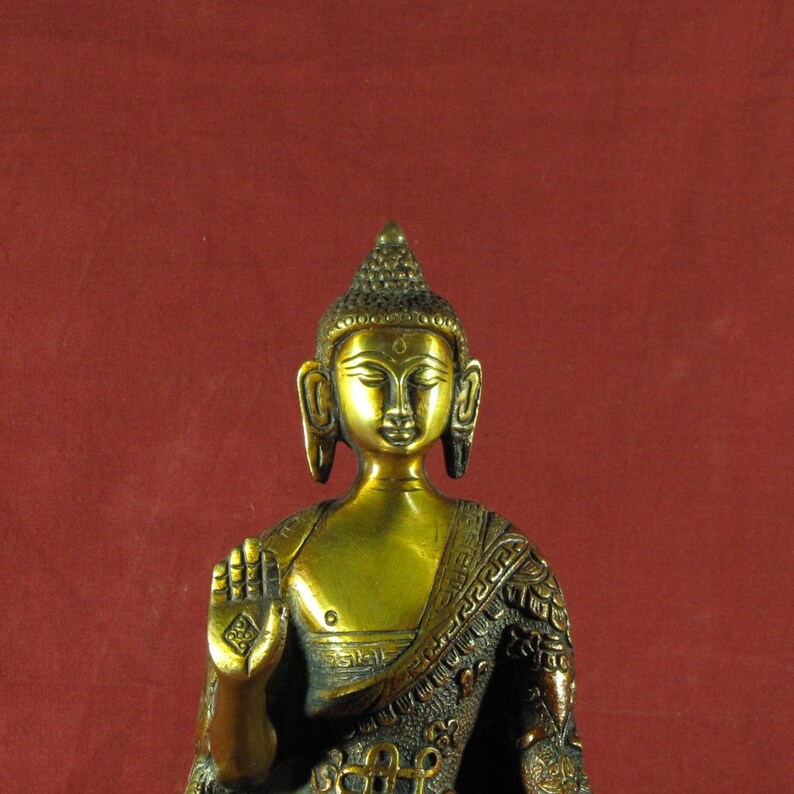 May include: A gold-coloured statue of Buddha with a serene expression. The statue is sitting with one hand raised in a gesture of blessing. The statue is wearing a robe with intricate details.