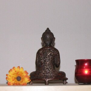 May include: A dark brown, ornate, metal statue of a seated Buddha. The statue is on a small, decorative base with three legs. The Buddha is facing forward with its hands in its lap.
