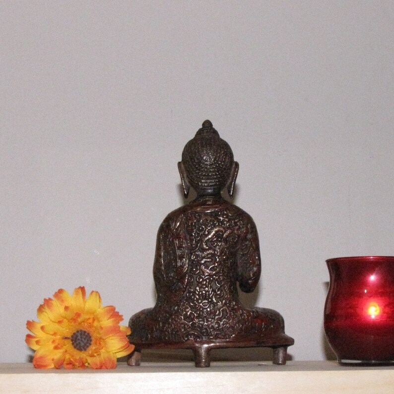 May include: A dark brown, ornate, metal statue of a seated Buddha. The statue is on a small, decorative base with three legs. The Buddha is facing forward with its hands in its lap.