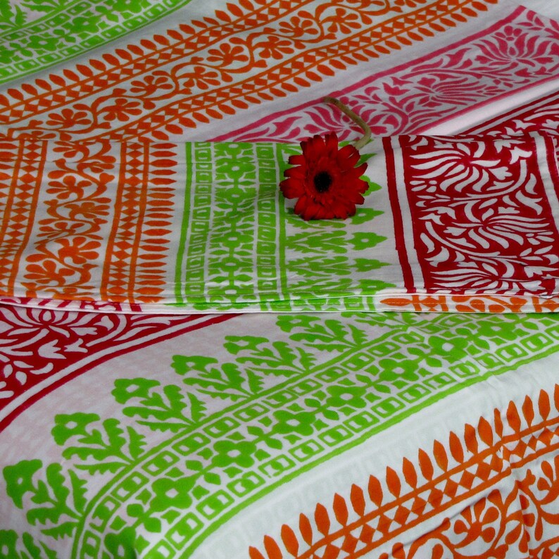 Pu&ograve; includere: Un tessuto bianco con strisce colorate di verde, arancione e rosa. Il tessuto presenta un motivo floreale ripetitivo. Un solo fiore rosa &egrave; posto sopra il tessuto.