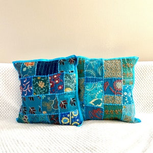 May include: Two square patchwork cushions in teal. Each cushion features a patchwork design with squares of teal, blue, gold, and red fabrics. They have bright blue borders. The cushions are placed on a white surface.