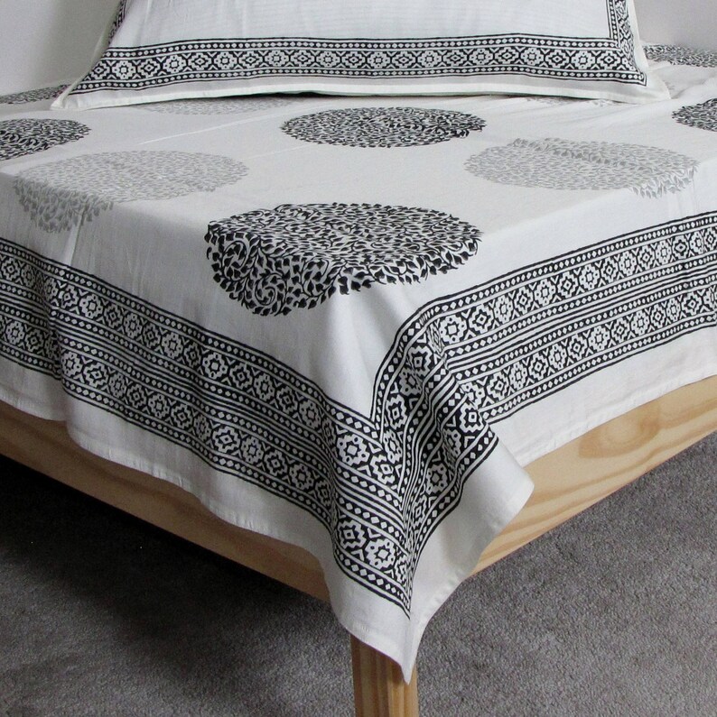 May include: White cotton bed sheet with a black and white geometric pattern. The sheet has a border of repeating geometric designs and a central design of circles with a floral pattern.