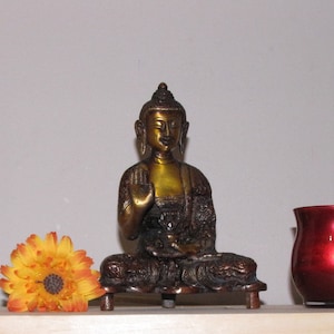 May include: A gold-colored metal statue of Buddha sitting in a meditative pose. The statue is on a wooden base with a flower in the foreground.