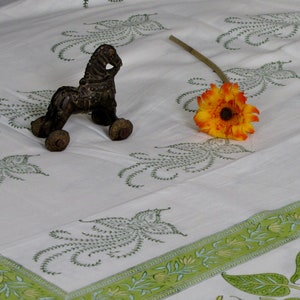 May include: A white cotton tablecloth with a green floral pattern. A small, dark brown metal toy horse is on the tablecloth, and a bright orange daisy is on the right side of the image.