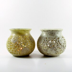 May include: Two white carved stone candle holders with intricate floral designs. The candle holders are round and have a wide opening at the top. The candle holders are made of soapstone and have a natural, rustic look.