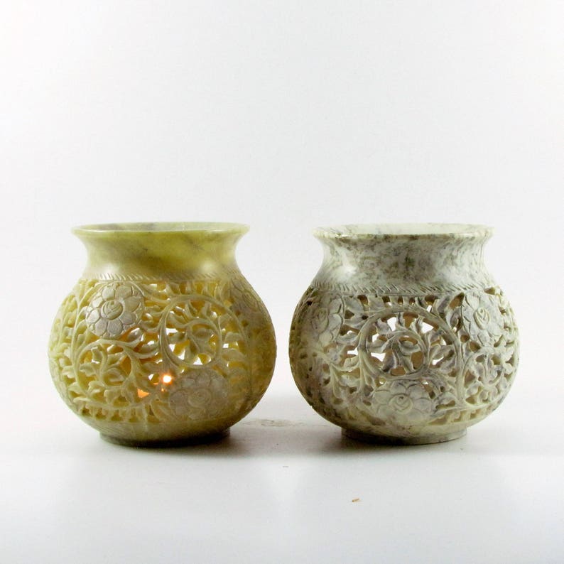 May include: Two white carved stone candle holders with intricate floral designs. The candle holders are round and have a wide opening at the top. The candle holders are made of soapstone and have a natural, rustic look.