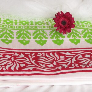 Pu&ograve; includere: Un tessuto di cotone bianco con una stampa floreale verde e rossa. Il tessuto ha un bordo con un motivo ripetitivo di foglie e fiori. Un fiore rosa &egrave; posto sopra il tessuto.