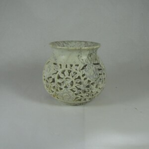 May include: A white marble candle holder with intricate floral carvings. The holder has a round base and a wide, open top.
