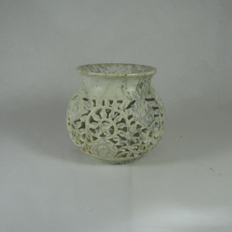 May include: A white marble candle holder with intricate floral carvings. The holder has a round base and a wide, open top.