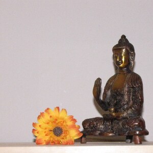 May include: A golden bronze statue of a seated Buddha with a raised hand, on a wooden base. A yellow and orange flower is in the foreground.