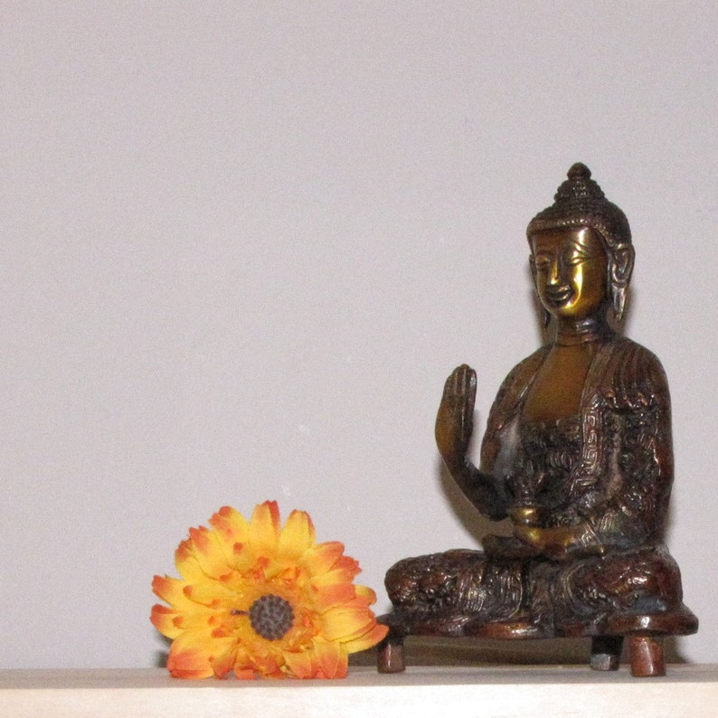 May include: A golden bronze statue of a seated Buddha with a raised hand, on a wooden base. A yellow and orange flower is in the foreground.