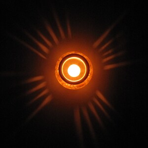 May include: A round, orange candle holder with a white candle inside. The candle holder casts a warm glow and creates a starburst pattern of light on the dark background.