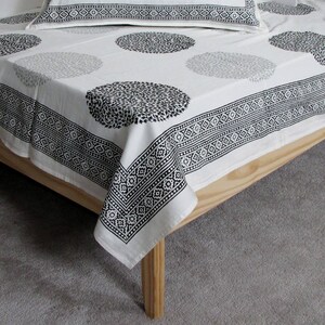 May include: White cotton bed sheet with a black and white geometric border and a repeating pattern of black circles with a floral design.