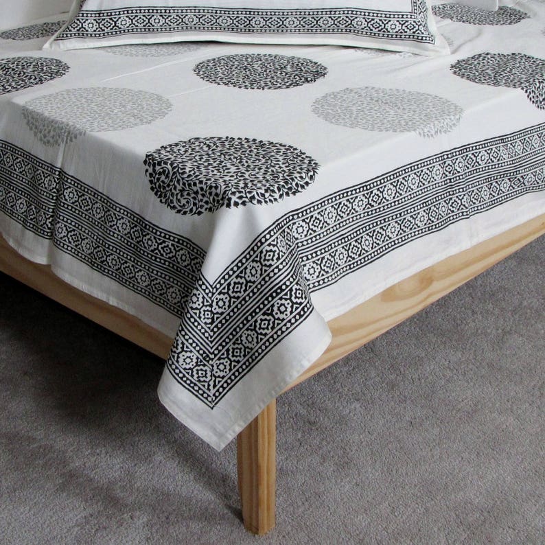 May include: White cotton bed sheet with a black and white geometric border and a repeating pattern of black circles with a floral design.
