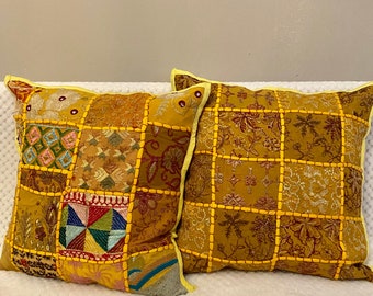 Può includere: Due cuscini decorativi con un design a patchwork. I cuscini sono realizzati in tessuto giallo con ricami marroni e rossi. I cuscini hanno un bordo giallo.