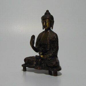 May include: A small, gold-toned bronze statue of a seated Buddha. The Buddha is in a meditative pose with one hand raised in a gesture of blessing. The statue is on a small, rectangular base.
