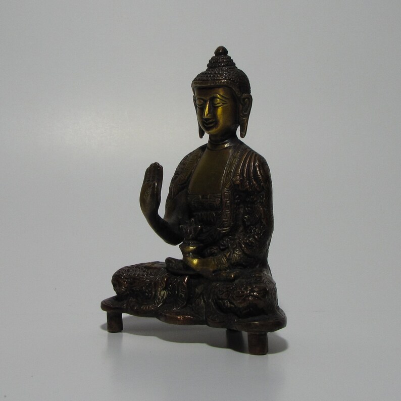 May include: A small, gold-toned bronze statue of a seated Buddha. The Buddha is in a meditative pose with one hand raised in a gesture of blessing. The statue is on a small, rectangular base.