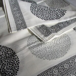 May include: White cotton fabric with a black and gray floral pattern. The fabric is folded in thirds, with the top layer folded over the bottom two layers. The fabric has a border of black and white geometric designs.