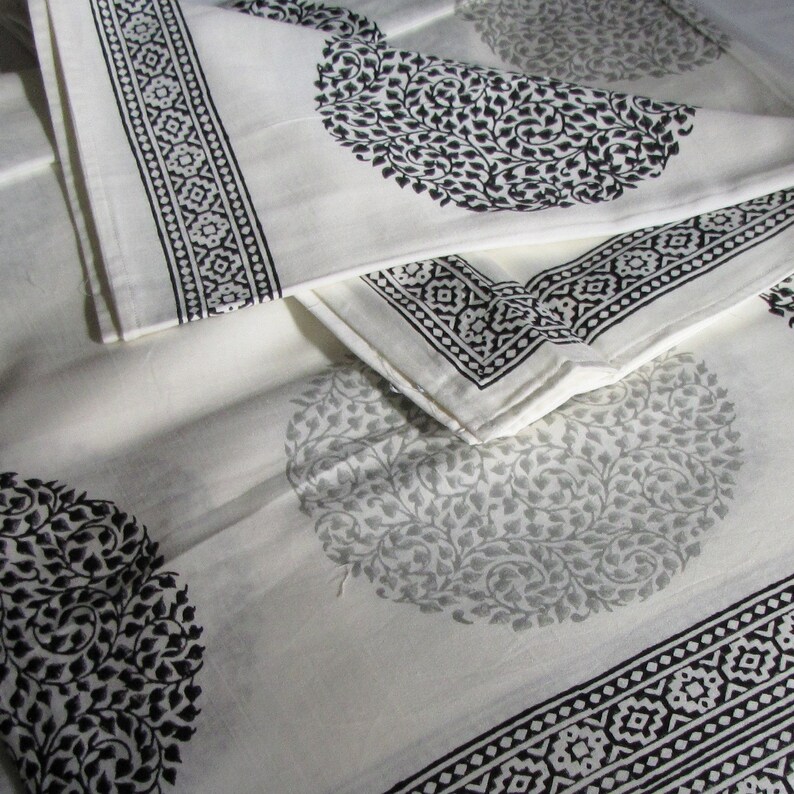 May include: White cotton fabric with a black and gray floral pattern. The fabric is folded in thirds, with the top layer folded over the bottom two layers. The fabric has a border of black and white geometric designs.
