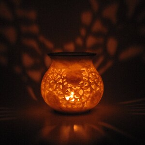 May include: A white carved stone candle holder with intricate floral designs. A candle is lit inside the holder, casting a warm glow.