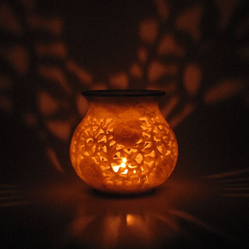 May include: A white carved stone candle holder with intricate floral designs. A candle is lit inside the holder, casting a warm glow.