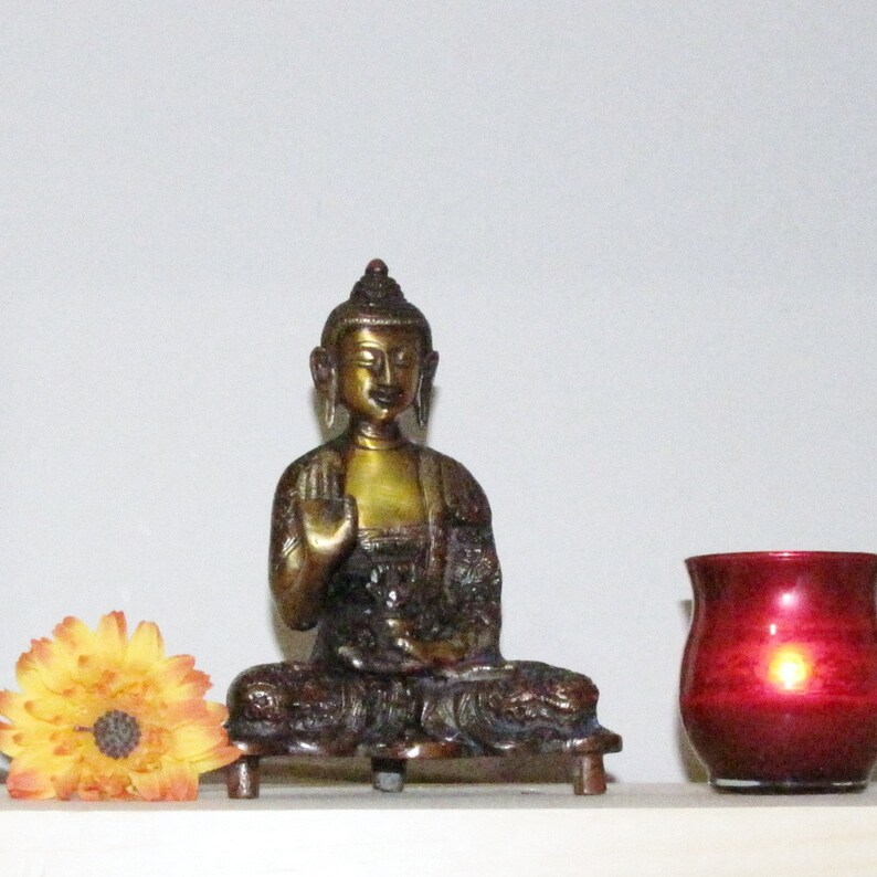 May include: A gold-toned metal statue of a seated Buddha with a serene expression. The statue is on a small base and has a red candle holder in the background.
