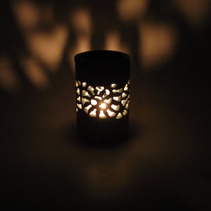 May include: A black ceramic oil burner with a decorative carved design. The burner has a small candle inside, casting a warm glow through the intricate carvings.