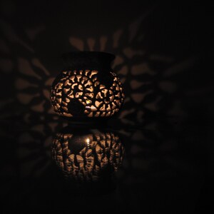 May include: A white ceramic candle holder with intricate floral designs. The candle holder is lit and casting shadows on the surface.