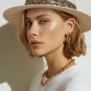 May include: A beige straw fedora hat featuring a black band with a geometric pattern of sequins in gold, pink, and green. The hat is paired with a gold chain link necklace and small gold hoop earrings, worn with a plain white crew neck shirt.