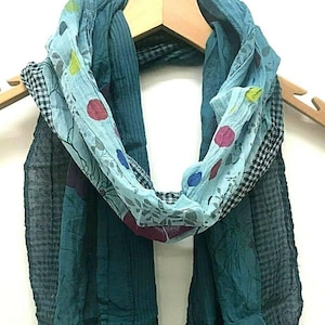 May include: A teal scarf with a variety of patterns and textures. The scarf features a light blue section with colorful polka dots and floral designs. The edges have a fringed detail. The scarf is hanging on a wooden hanger.