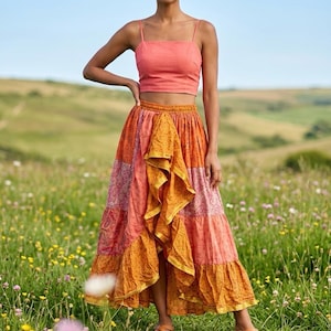 May include: A woman in a coral crop top and a long, patchwork skirt with orange and pink panels. The skirt features a ruffled, asymmetrical hem. She is standing in a field of wildflowers, wearing brown sandals.
