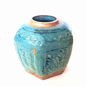 Puede incluir: Un tarro de cerámica de forma cuadrada con un esmalte turquesa vibrante y una amplia abertura circular. El exterior presenta patrones florales en relieve en cada lado, con un borde y base de arcilla beige sin esmaltar.