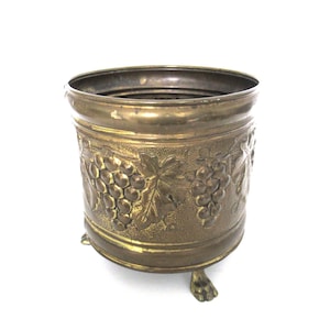 Könnte beinhalten: Ein Blumentopf aus Messing mit einem Traubenmuster und drei Füßen. Der Topf ist hellgoldfarben und hat eine abgenutzte Oberfläche.