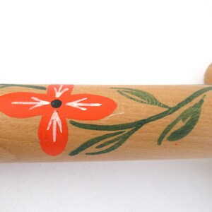 Puede incluir: Un rodillo de madera con un dise&ntilde;o pintado a mano de hojas verdes y una flor roja con un centro negro.