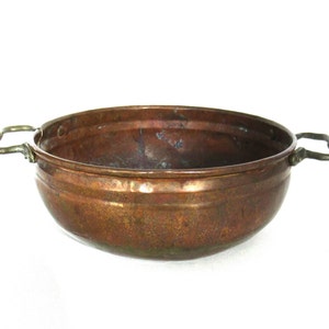 Op de afbeelding: Een koperen kom met twee messing handvatten. De kom heeft een ronde vorm en een licht gehammerde textuur.