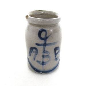 May include: A small, grey stoneware crock with a blue anchor design. The anchor is hand-painted and has a simple, rustic style.