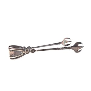May include: Antique silver sugar tongs with a floral design on the handle. The tongs have a decorative, curved handle and two claw-like ends for gripping sugar cubes. The silver has a tarnished appearance, suggesting age and use.