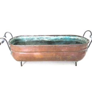 Può includere: Una fioriera rettangolare color rame con una patina invecchiata. Presenta manici curvi con dettagli in ceramica bianca e blu. La fioriera poggia su piccoli piedini in metallo.
