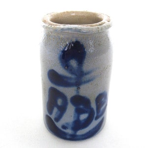 May include: A small, grey stoneware jar with a blue glaze. The jar features a stylised design of a flower or plant on the front.