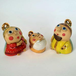 以下が含まれることがあります： 奇抜な顔をした3つのセラミックフィギュア。フィギュアは大きな青い目、バラ色の頬、金色のアクセントが特徴です。あるフィギュアは赤いドレス、別のフィギュアは黄色のコート、3つ目は白いシャツを着ています。フィギュアは白い背景にあります。
