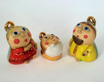 Puede incluir: Tres figuras de cerámica con caras caprichosas. Las figuras tienen grandes ojos azules, mejillas rosadas y detalles dorados. Una figura lleva un vestido rojo, otra un abrigo amarillo y la tercera una camisa blanca. Las figuras están sobre un fondo blanco.
