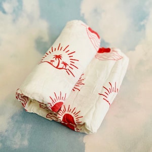 Puede incluir: Dos arrullos de muselina blanca con diseños rojos de palmeras y soles. Los arrullos están doblados y apilados uno encima del otro.